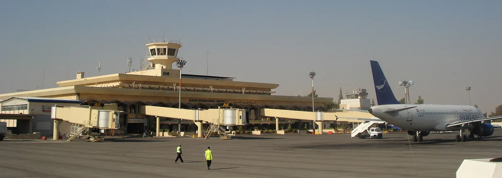 Low cream terminal building with a square control tower, seen across the apron with a Syrianair airliner parked on the right under a pale sky