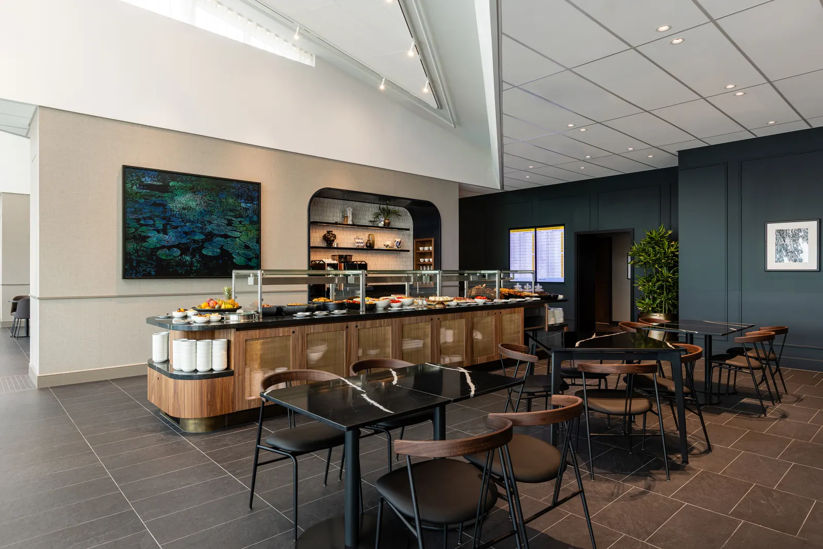 Dining tables with black marble tops beside a wood-paneled buffet stocked with fruit and plated dishes inside the American Express Centurion Lounge at Amsterdam Schiphol (AMS).