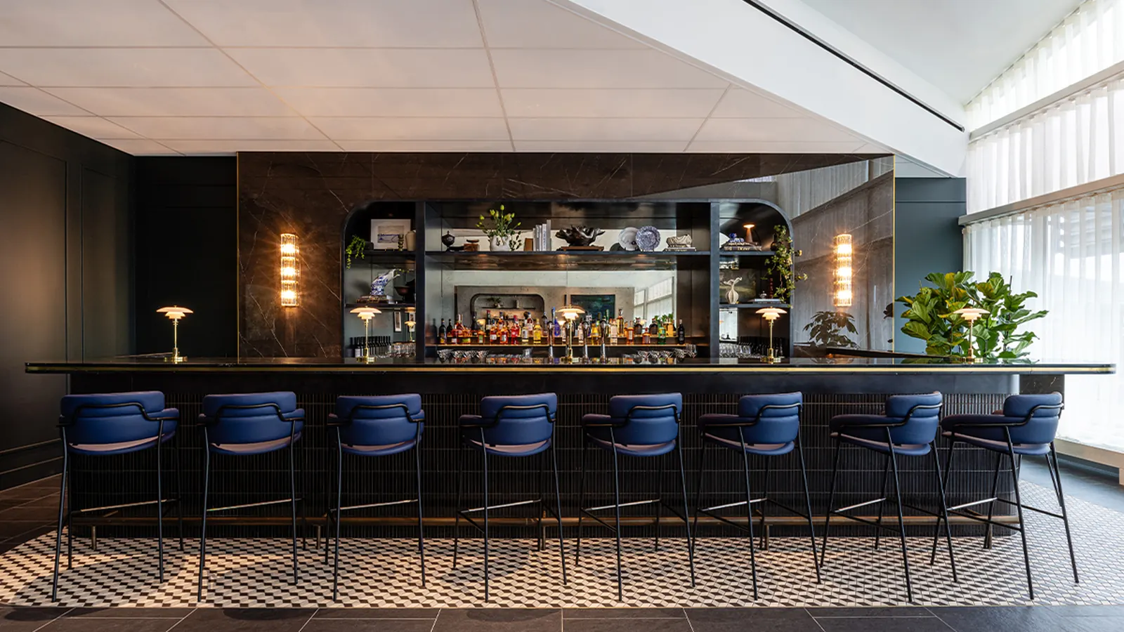 Long dark marble bar with navy blue stools, brass table lamps, and a mirrored back bar inside the American Express Centurion Lounge at Amsterdam Schiphol (AMS).