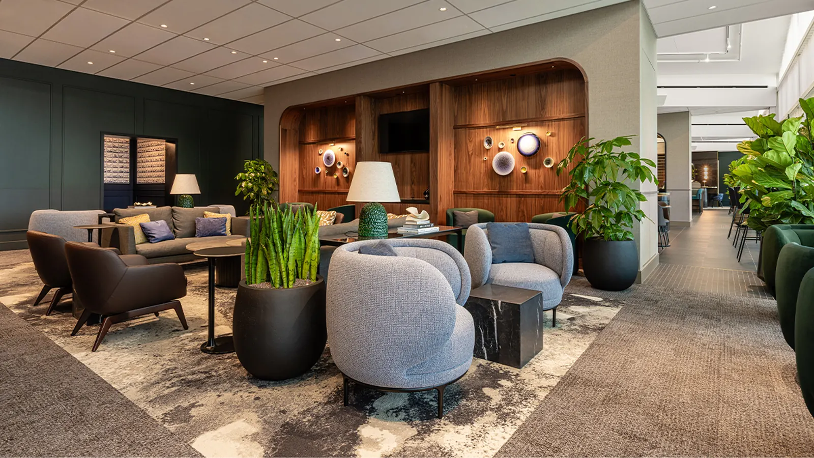 Grey fabric armchairs, wood-paneled TV alcove with Delft-blue plate art, and planters in a plush seating cluster inside the American Express Centurion Lounge at Amsterdam Schiphol (AMS).