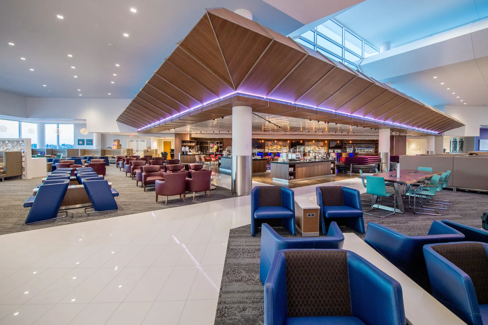 Wood canopy over a central buffet with blue and burgundy lounge chairs and a teal communal table inside the Delta Sky Club on Concourse B at Hartsfield-Jackson Atlanta (ATL).