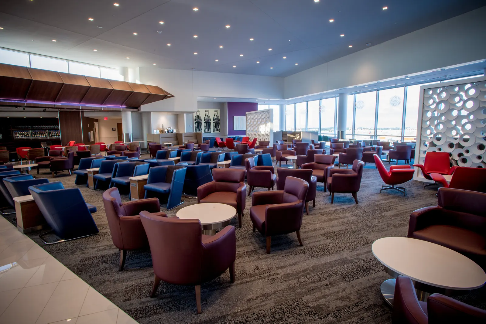 Burgundy and navy armchairs with powered side tables, circular white partitions, and floor-to-ceiling apron windows inside the Delta Sky Club on Concourse B at Hartsfield-Jackson Atlanta (ATL).