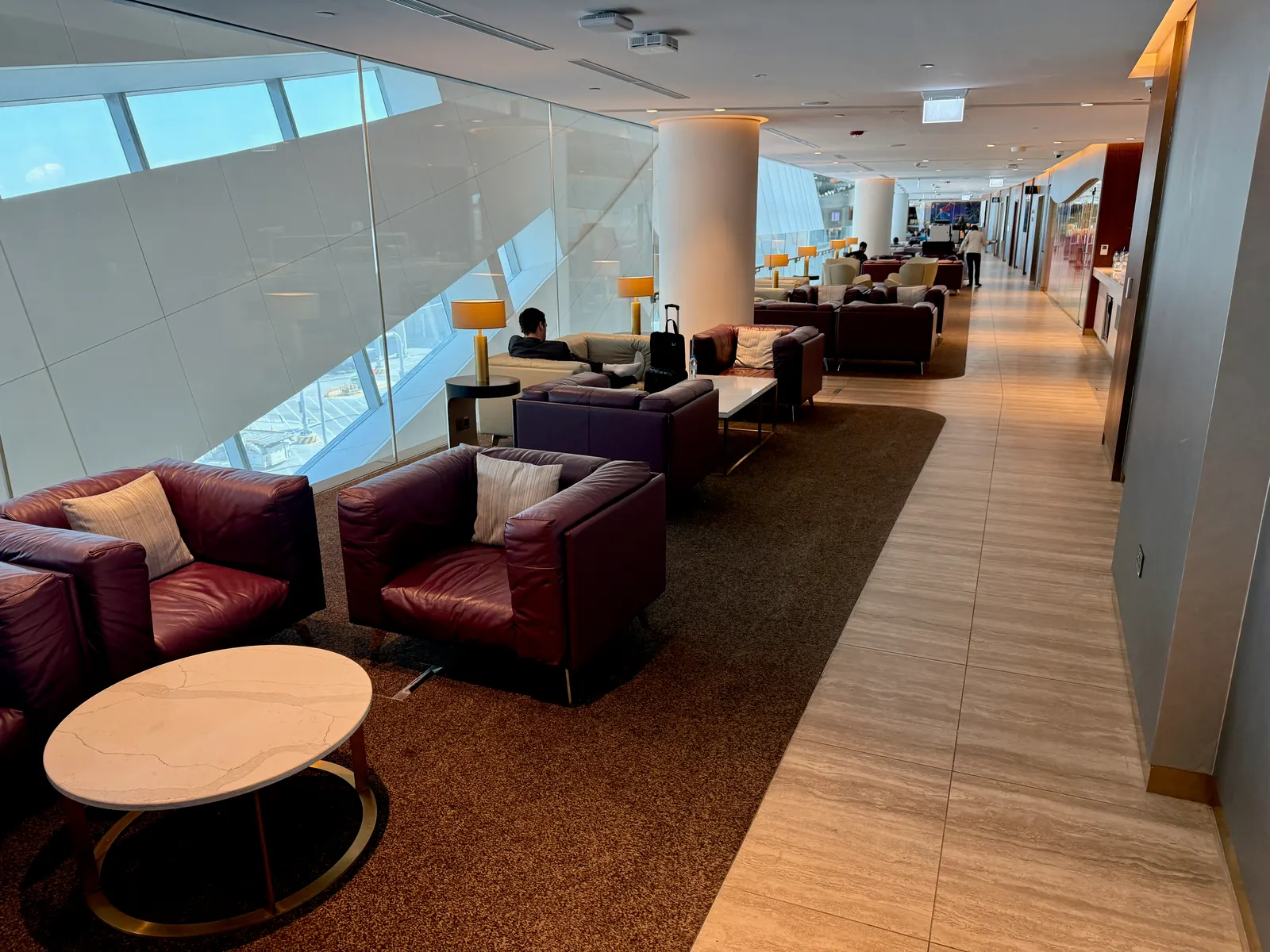 Purple leather armchairs, marble coffee tables, and warm gold lamps along a window wall inside the Etihad Business Class Lounge at Abu Dhabi International (AUH).