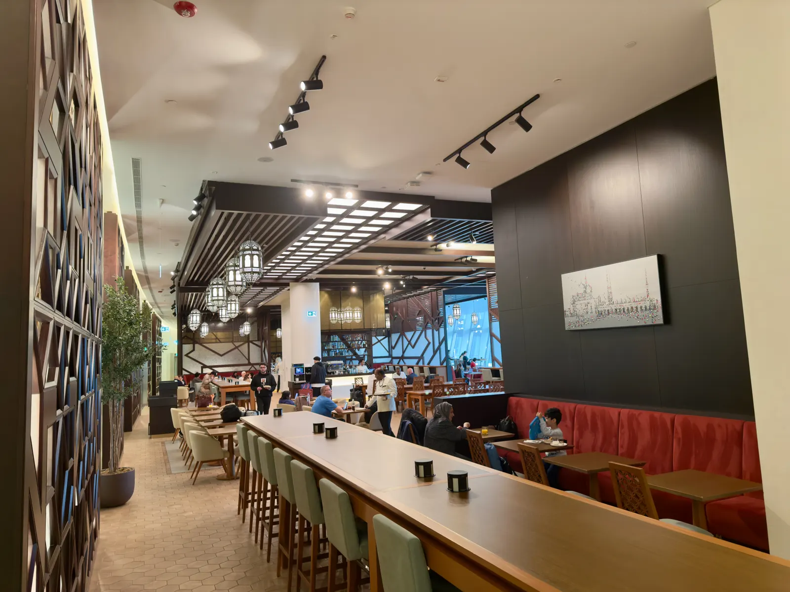 Long communal wood table with green stools, red banquettes, and geometric lantern lights inside the Etihad Business Class Lounge at Abu Dhabi International (AUH).