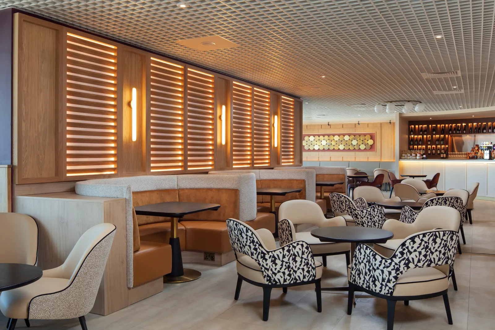 Cream curved banquettes, dalmatian-print armchairs and black round tables against wood-slat lighting in Luxe by Aspire at Birmingham Airport (BHX).