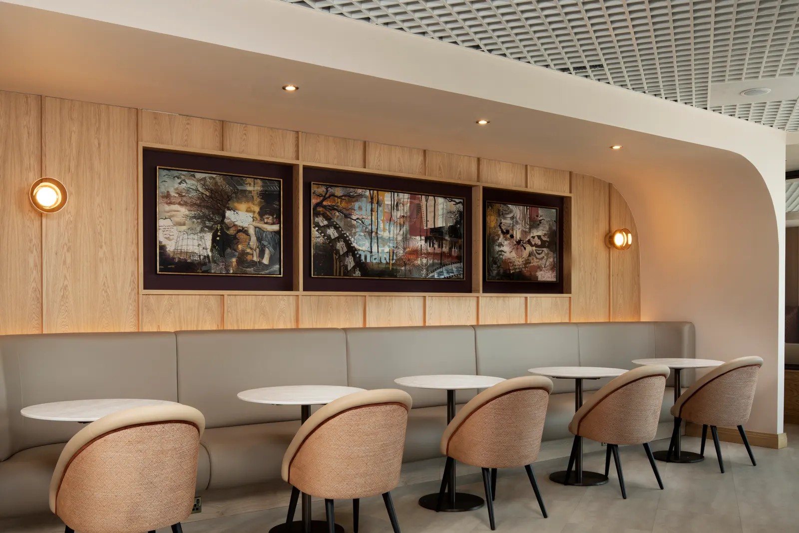 Long grey banquette under framed landscape art with tan armchairs and white pedestal tables in Luxe by Aspire at Birmingham Airport (BHX).