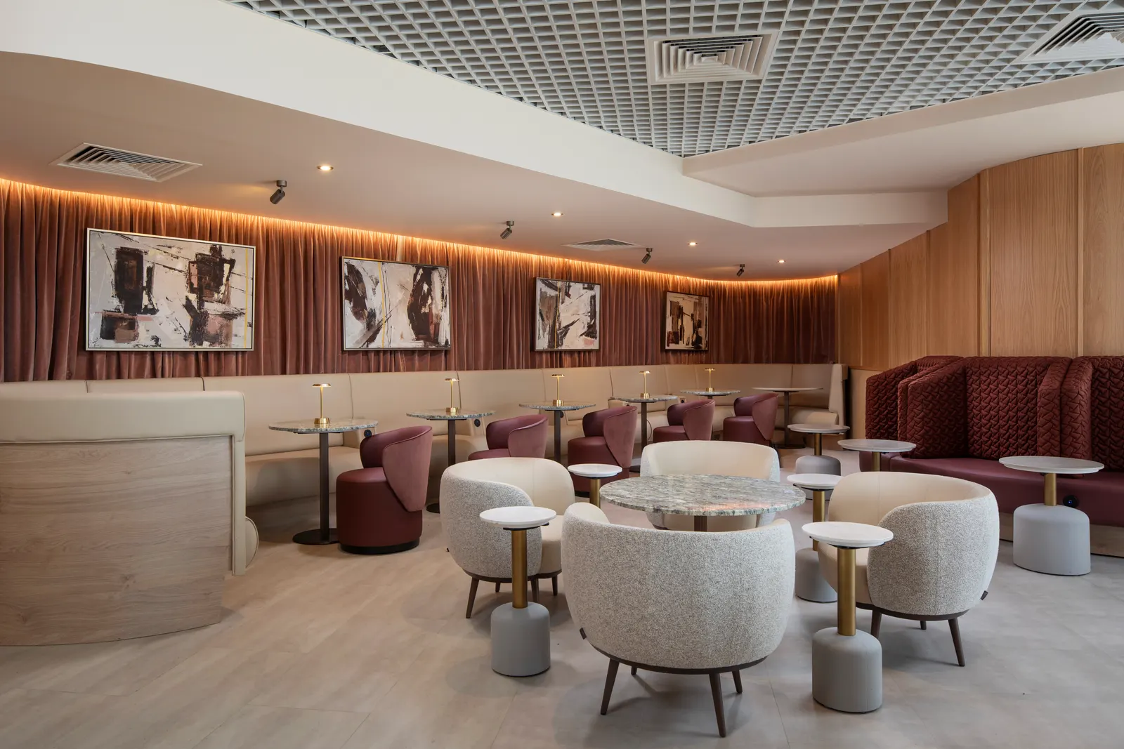 Clusters of cream armchairs around marble tables with a maroon banquette and pink vertical slats in Suite by Aspire at Birmingham Airport (BHX).
