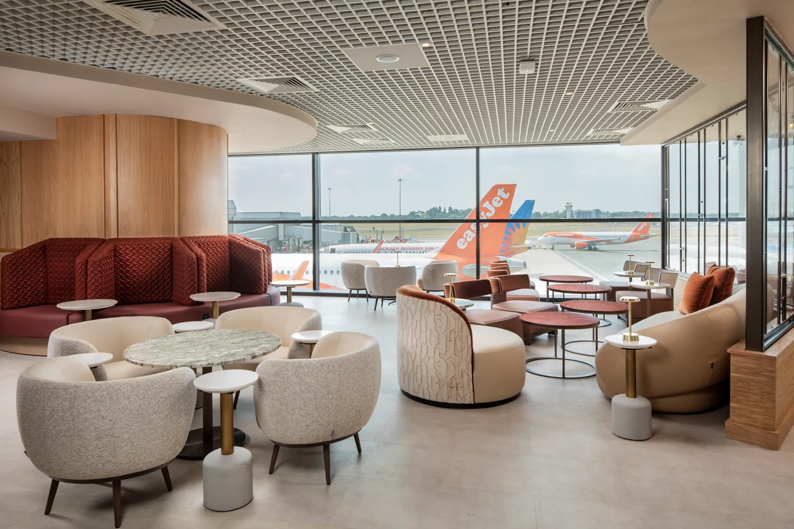 Cream armchairs and maroon banquettes beside floor-to-ceiling apron windows in Suite by Aspire at Birmingham Airport (BHX).