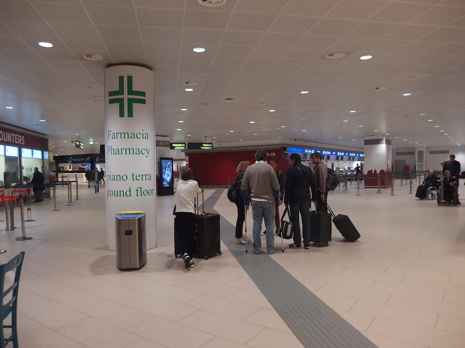 Check-in hall at Bologna Guglielmo Marconi Airport with pharmacy column, passengers and check-in belts