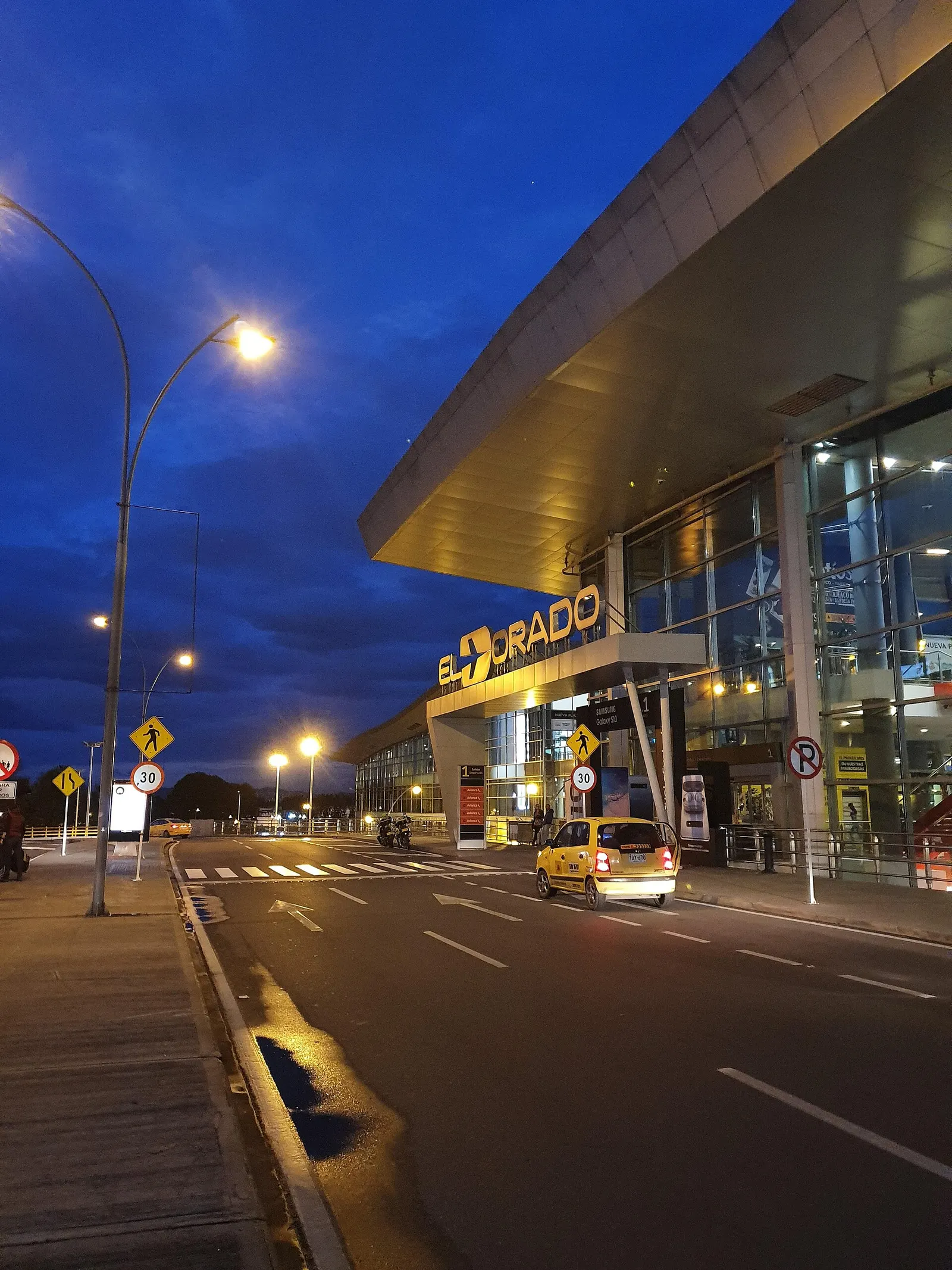 The illuminated EL DORADO sign and glass-fronted main terminal facade at night, seen from the airport access road with a taxi crossing the foreground.