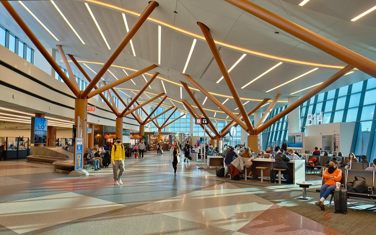 Sunlit Terminal B concourse at Boston Logan near gate B14, with branched wood columns, seating and shops.
