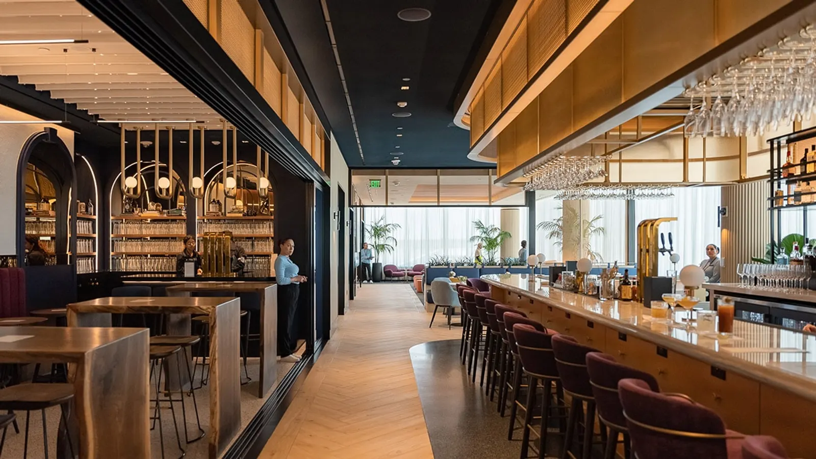 Long wood bar with burgundy stools, arched back-bar shelving and hanging glassware in the Chase Sapphire Lounge by The Club at Boston Logan (BOS).