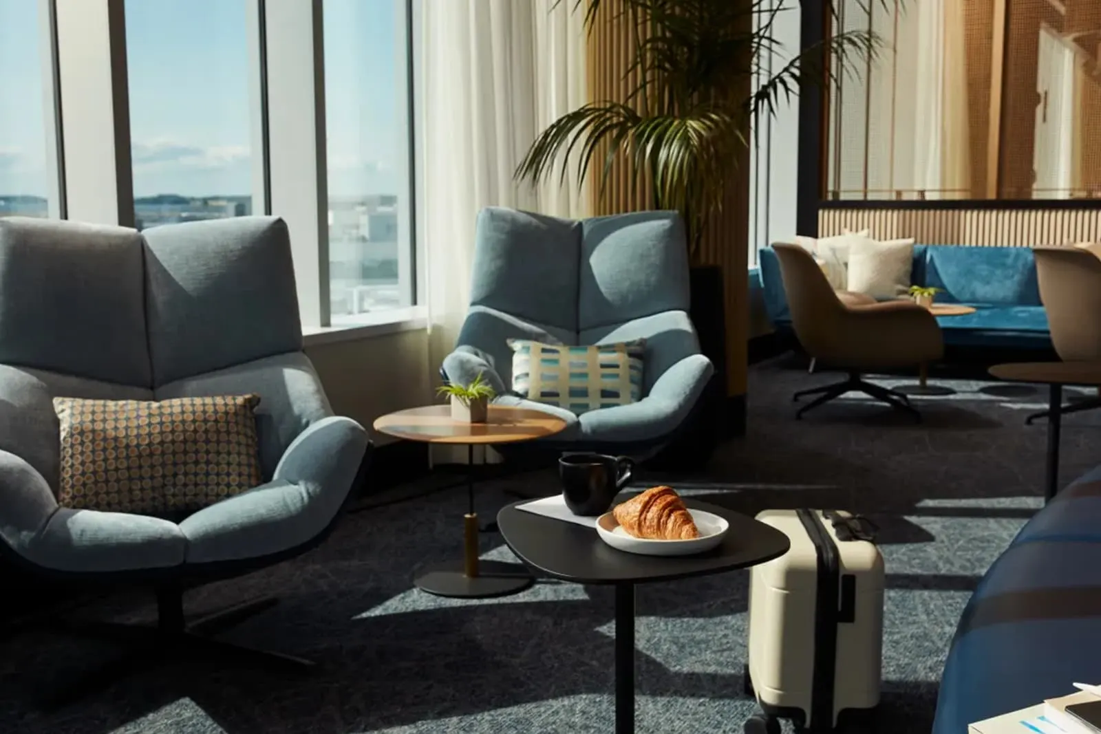 Blue-grey armchairs around small tables beside windows and potted palms in the Chase Sapphire Lounge by The Club at Boston Logan (BOS).