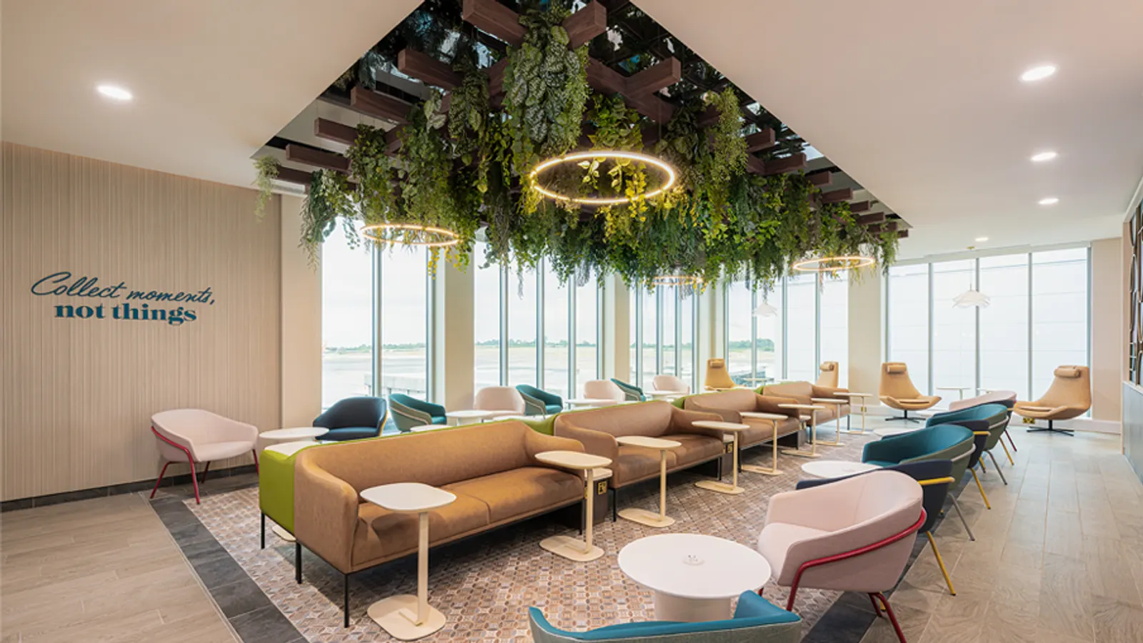 Green and tan sofas with pink armchairs under a plant-filled wooden lattice ceiling overlooking the apron in the Escape Lounge at Bristol Airport (BRS).