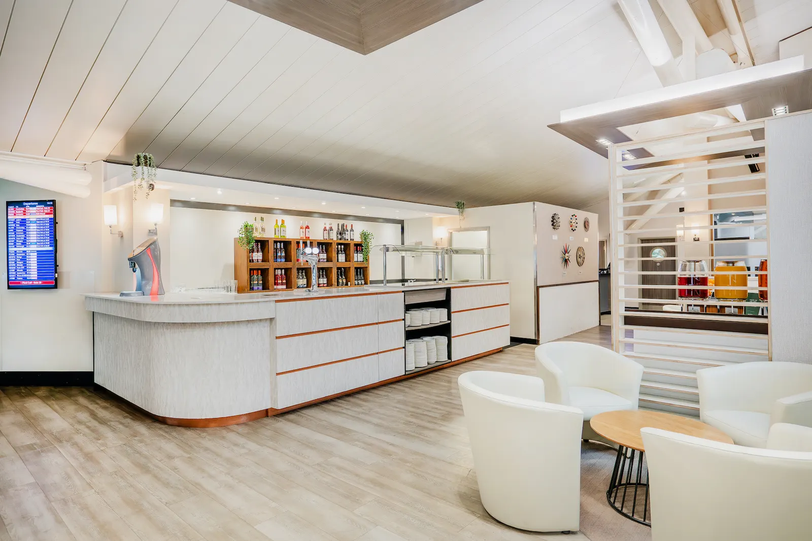White service counter with bottle shelving and white tub chairs around a wood table in Essence by Escape Lounges at Bristol Airport (BRS).