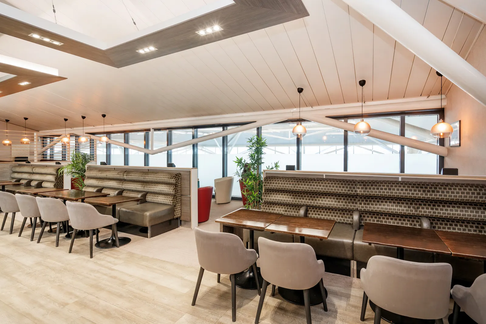 Dark banquette and grey armchairs along a light-wood floor under slanted white beams in Essence by Escape Lounges at Bristol Airport (BRS).