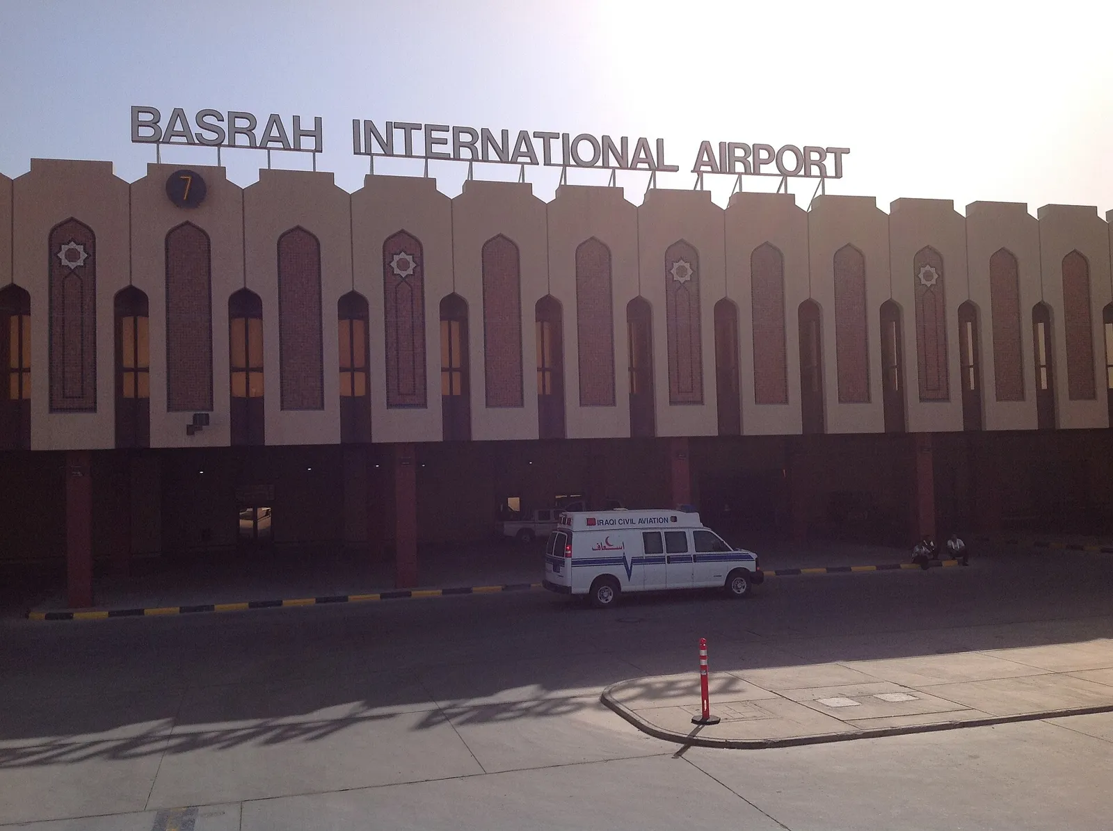 Sand-coloured terminal facade with arched windows and the BASRAH INTERNATIONAL AIRPORT sign on the roofline, an ambulance parked in front