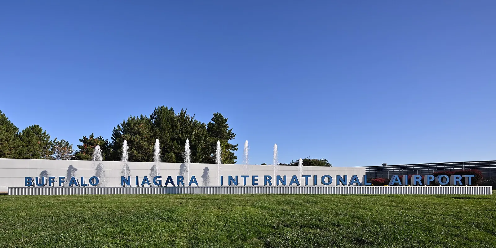 Blue three-dimensional letters spelling BUFFALO NIAGARA INTERNATIONAL AIRPORT in front of fountain jets and a white wall under a clear sky.