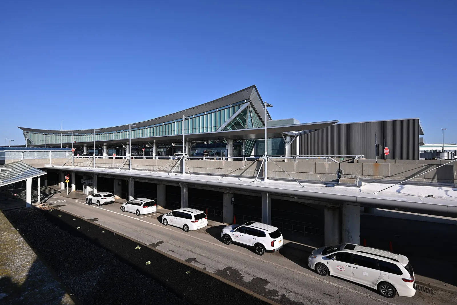 Curved glass BUF terminal with upper departures drop-off and lower arrivals curb, white vans waiting under the canopy on a clear September morning.