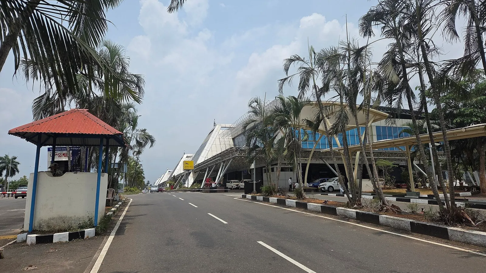 Landside kerb and sawtooth terminal façade at Calicut International Airport, with palms, a security hut, and the departures road.