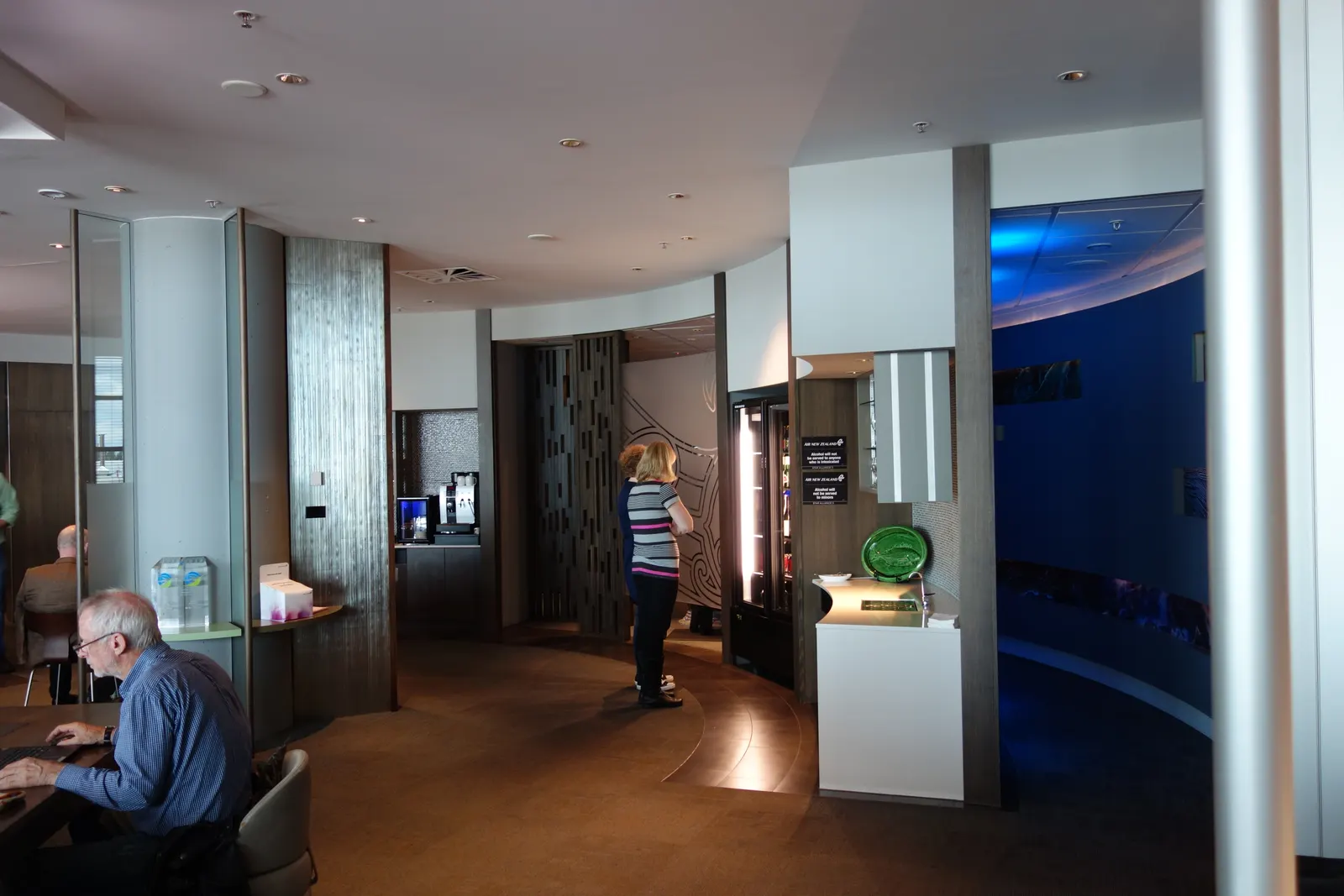 Curved white and metallic refreshment counters with a glass-door beverage fridge and espresso machine inside the Air New Zealand International Koru Lounge at Christchurch Airport (CHC).