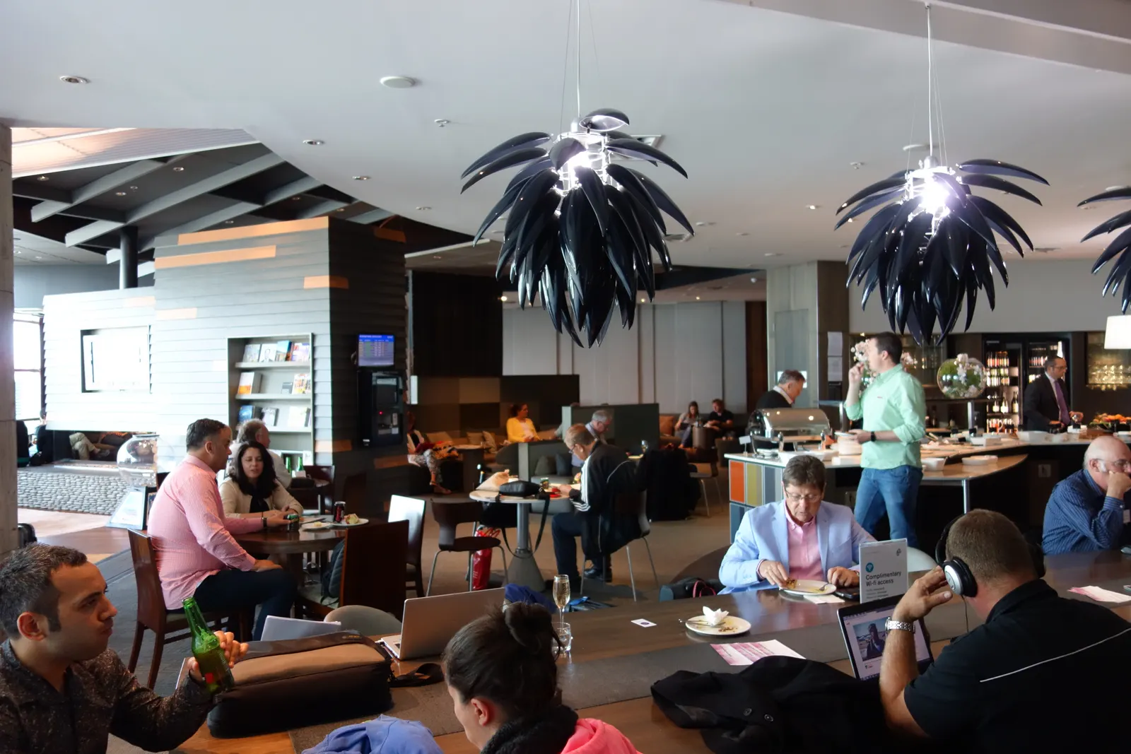 Communal work table and dining seating under black palm-frond chandeliers beside a buffet and flight information screen inside the Air New Zealand International Koru Lounge at Christchurch Airport (CHC).