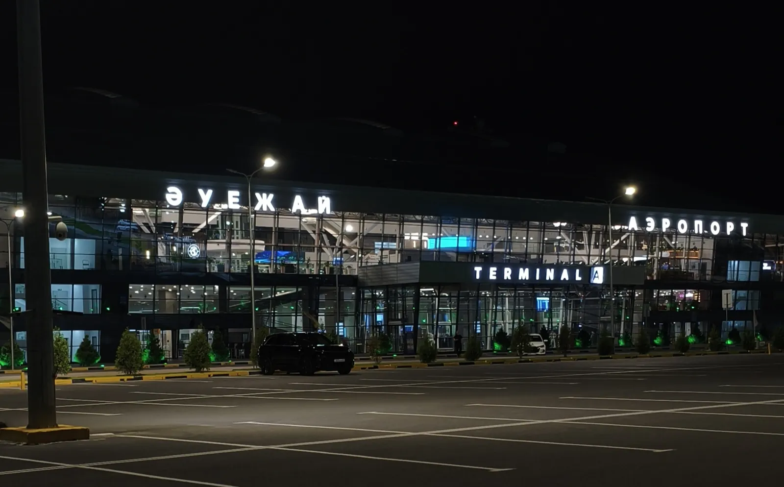 Glass-fronted Terminal A of Shymkent airport lit up at night, with 'Әуежай', 'Аэропорт' and 'Terminal A' signs on the roof and an almost empty forecourt