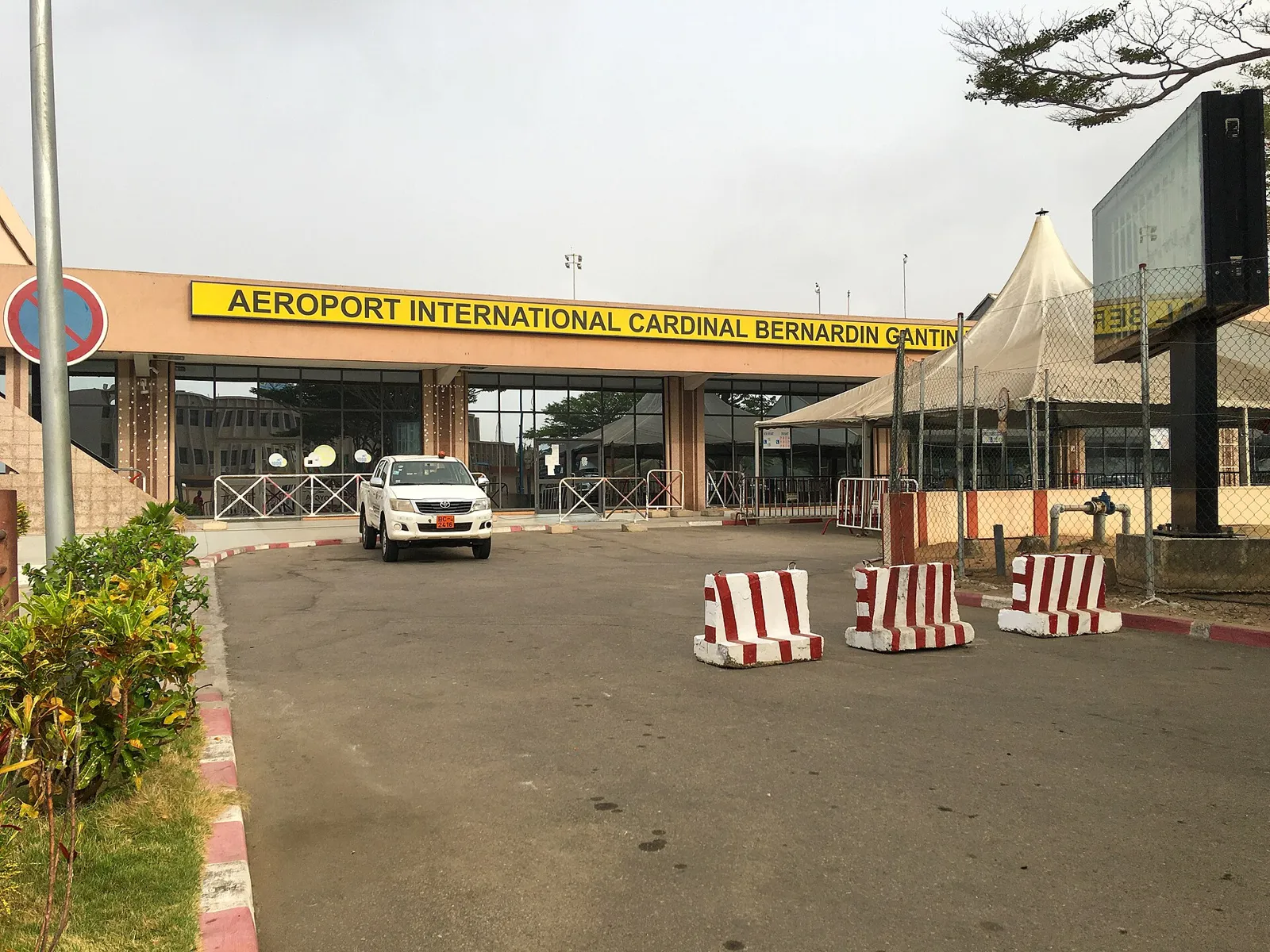 Single-storey terminal with a yellow sign reading Aeroport International Cardinal Bernardin Gantin above the entrance, red-and-white barriers on the forecourt