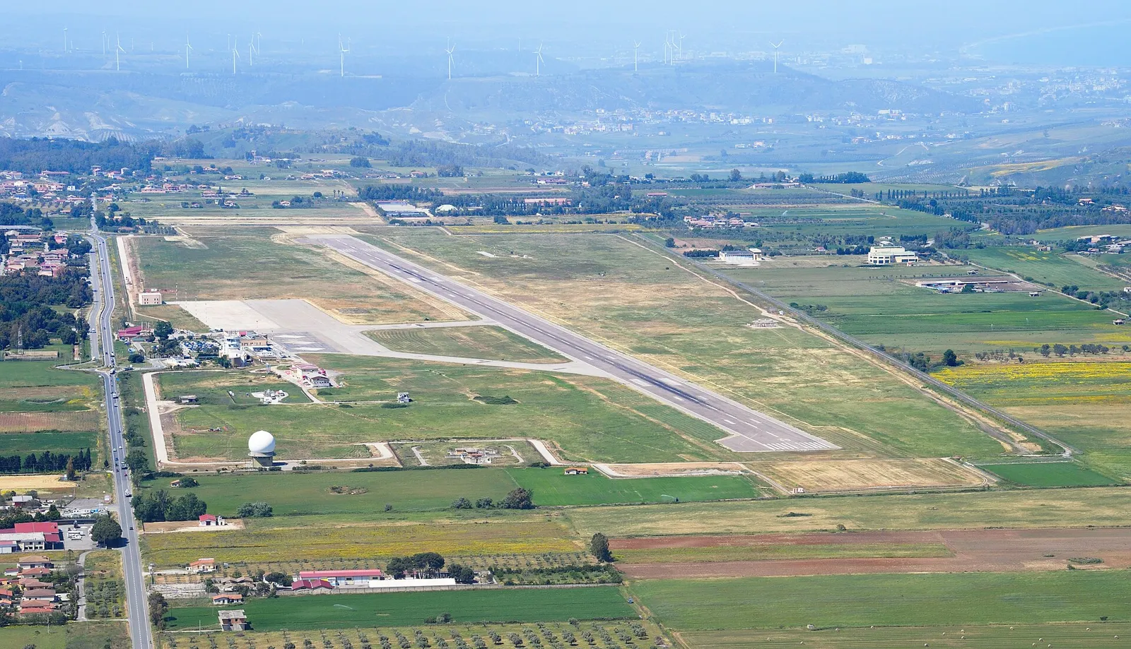 Aerial view of Crotone Airport's single runway and apron cutting across green farmland, with hills and a road along the left edge