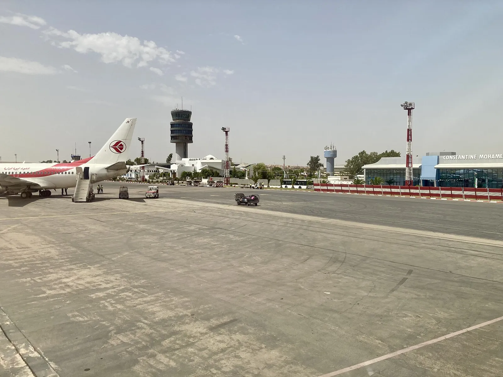 Air Algerie jet with boarding stairs on a wide concrete apron, the control tower in the middle distance and a glass terminal signed Constantine Mohamed Boudiaf on the right