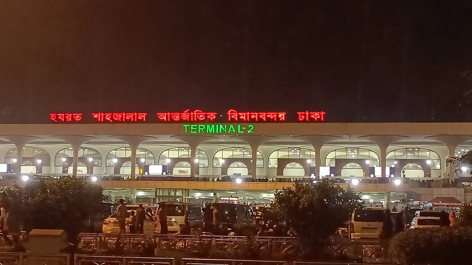 Night façade of Hazrat Shahjalal International Airport Terminal 2 in Dhaka, with the green TERMINAL-2 sign lit above the arrivals curb