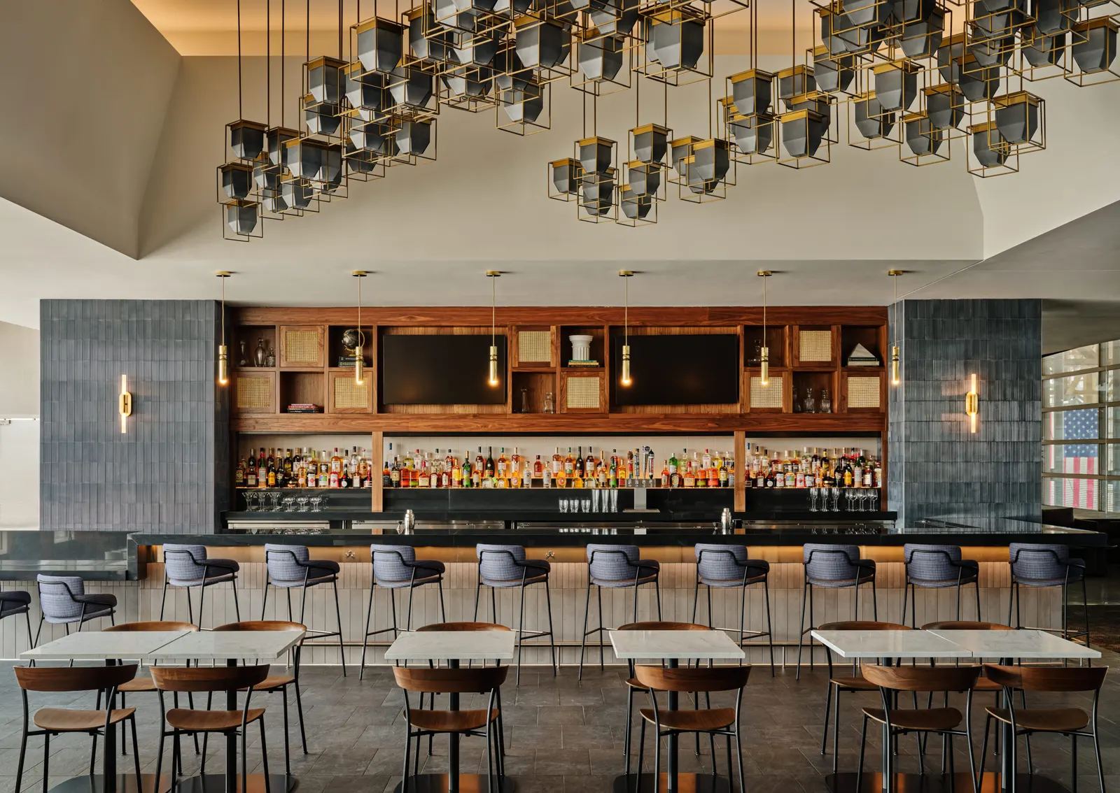 Wood-and-tile back bar with stools and communal dining tables beneath a hanging lantern sculpture in the Centurion Lounge at Ronald Reagan Washington National Airport (DCA).