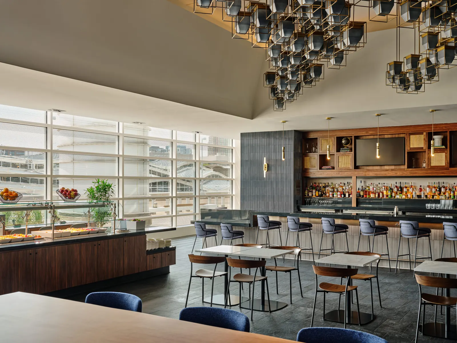 Dining tables and buffet counter under a lantern ceiling sculpture, with the bar and tall windows behind, in the Centurion Lounge at Ronald Reagan Washington National Airport (DCA).