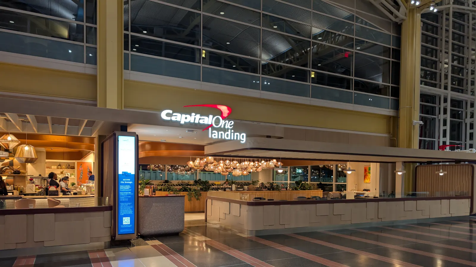 Night view into Capital One Landing showing the bar, dining tables, plant dividers, and clustered pendant lights at Ronald Reagan Washington National (DCA).