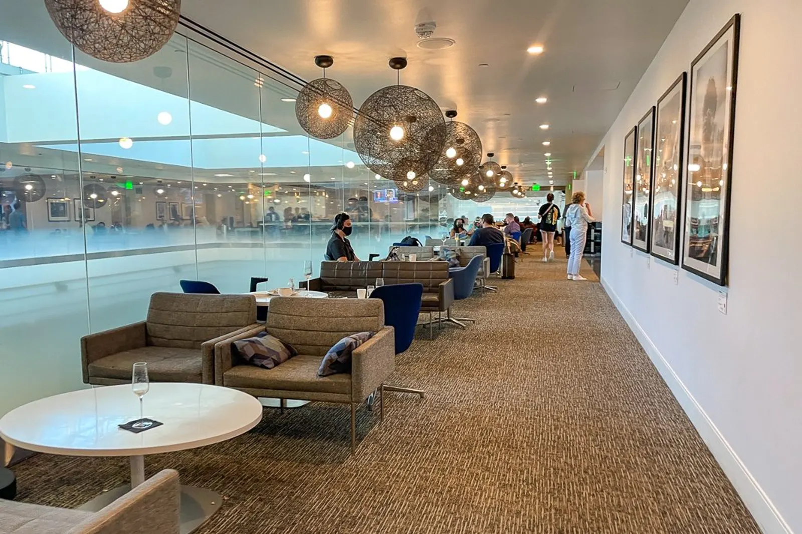 Long corridor of sofas and armchairs beside floor-to-ceiling glass overlooking Concourse C, under woven pendant lights, in the Centurion Lounge at Denver International Airport (DEN).