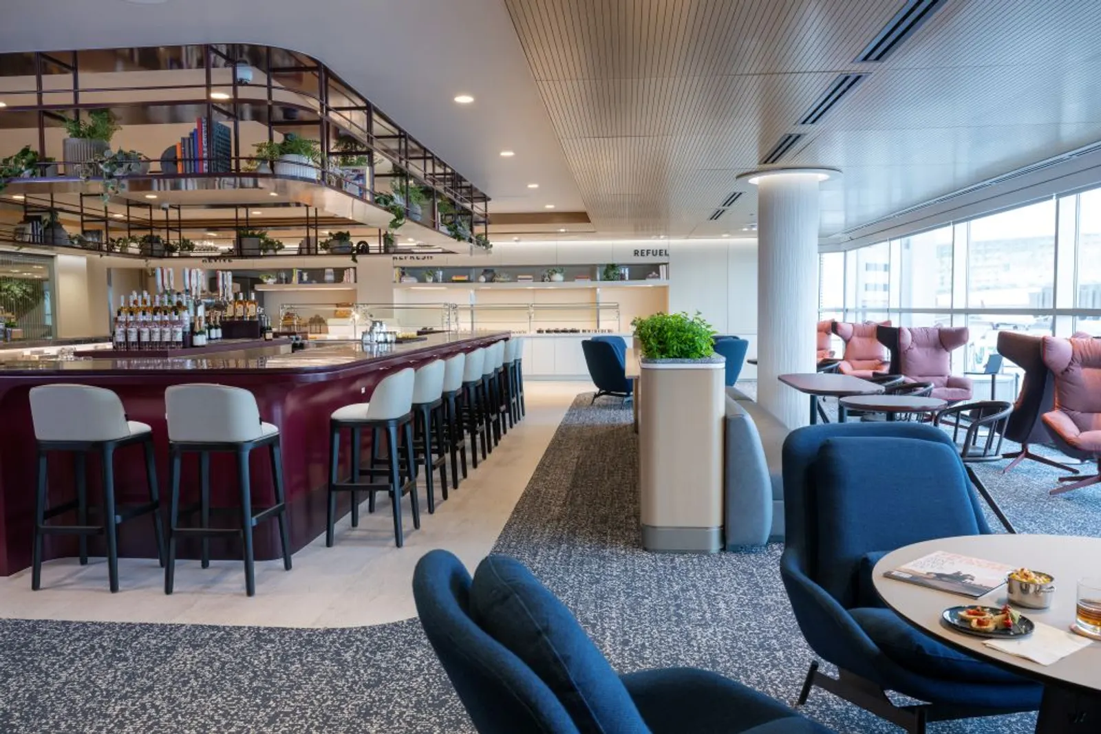 Burgundy-fronted full bar with white stools and an overhead shelving rack, beside blue armchair seating, in the Capital One Lounge at Denver International Airport (DEN).