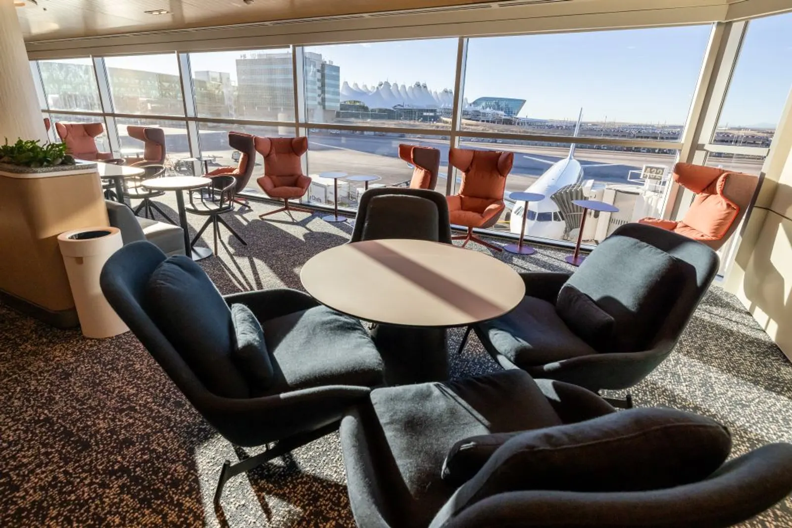 Black and coral armchairs around round tables beside floor-to-ceiling windows overlooking the apron, in the Capital One Lounge at Denver International Airport (DEN).