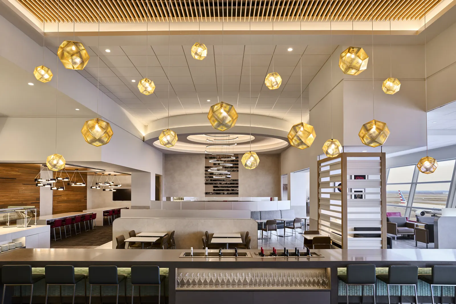 Bar counter and dining area under gold globe pendant lights and a wood-slat ceiling in the American Airlines Flagship Lounge at Dallas Fort Worth Terminal D (DFW).