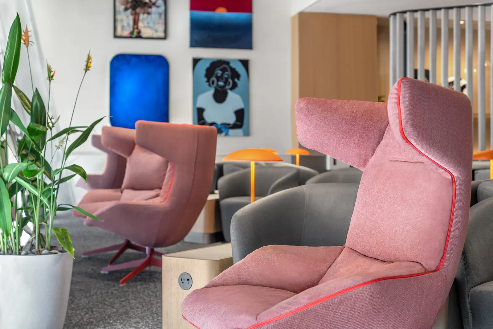 Pink high-back armchairs and grey seating beside potted plants and colorful local artwork in the Capital One Lounge at Dallas Fort Worth Terminal D (DFW).