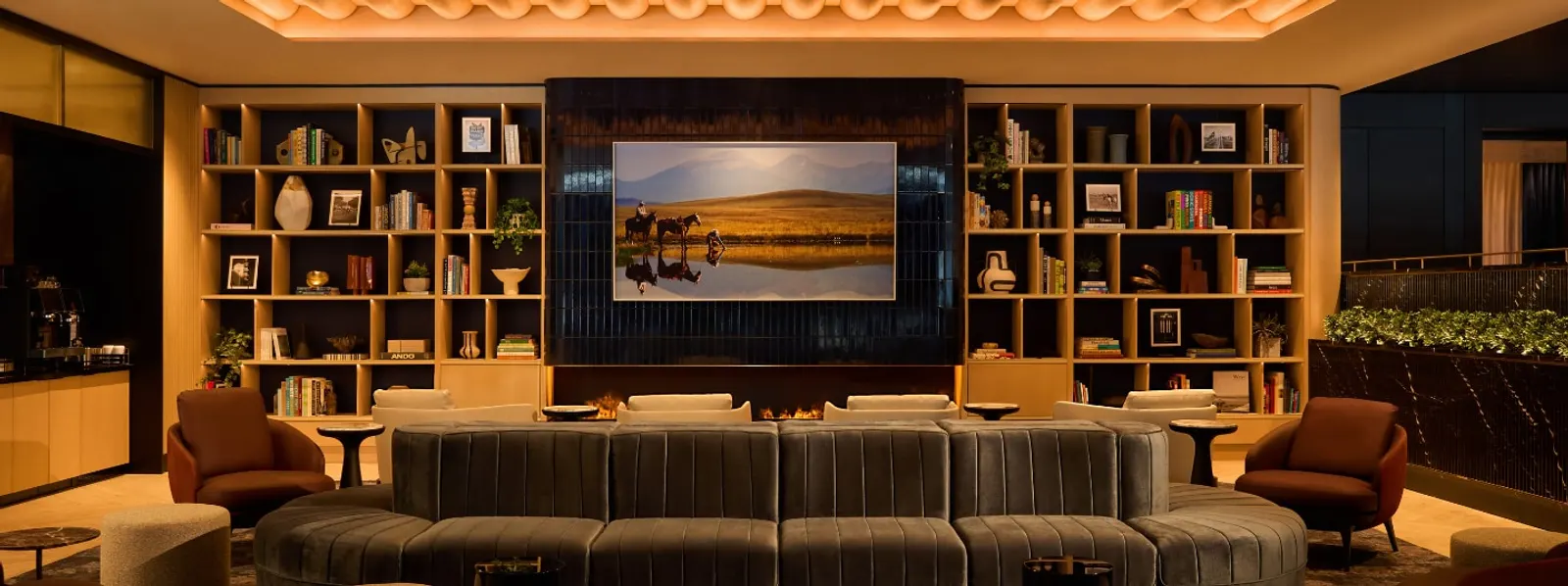 Living-room style seating with a long grey sofa and armchairs in front of wood bookshelves and a large landscape screen in the Chase Sapphire Lounge by The Club at Dallas Fort Worth (DFW).