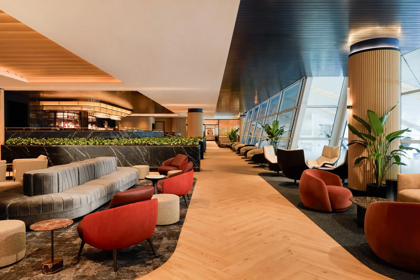 Red armchairs and grey sofas along a herringbone wood walkway beside floor-to-ceiling windows in the Chase Sapphire Lounge by The Club at Dallas Fort Worth Terminal D (DFW).