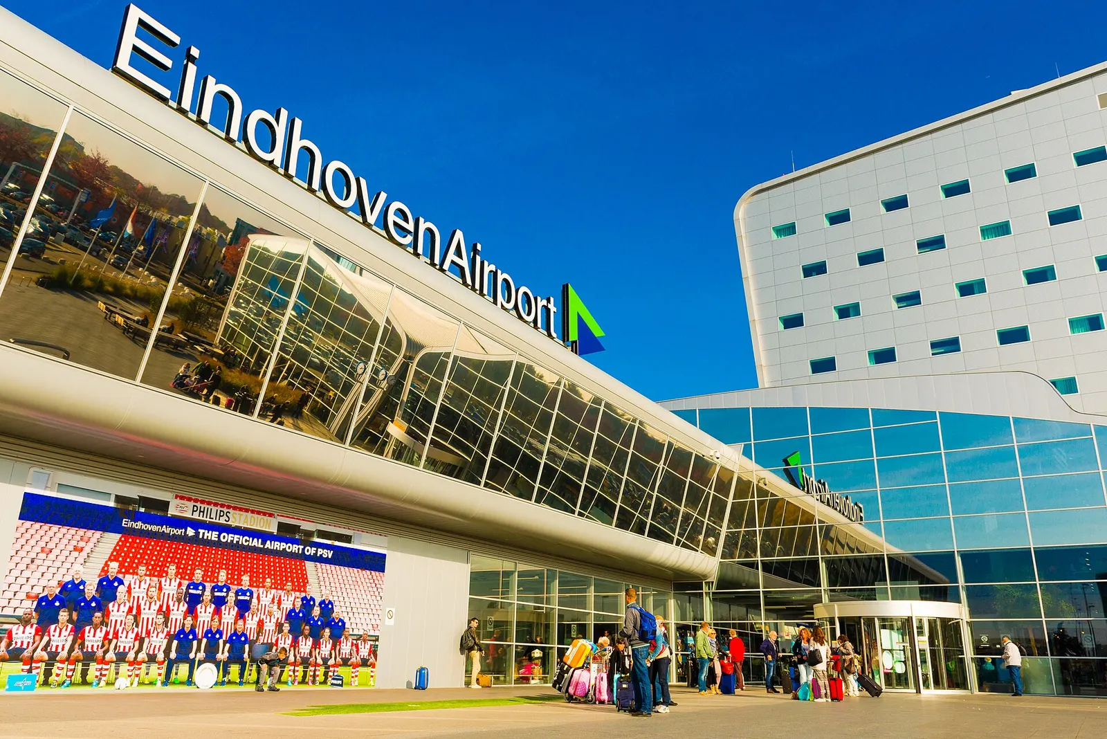 Front of the Eindhoven Airport terminal with the EindhovenAirport sign and passengers at the entrance