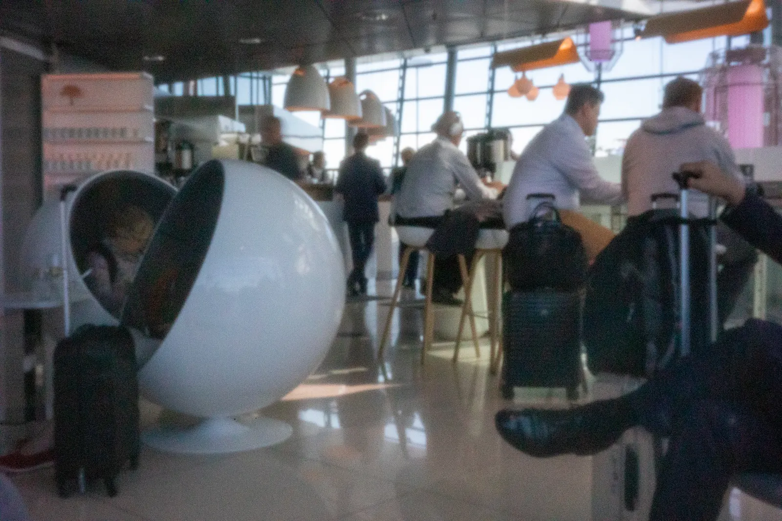 White spherical lounge chair beside black suitcases with bar stools and apron windows beyond inside the Finnair Lounge at Helsinki Airport (HEL).