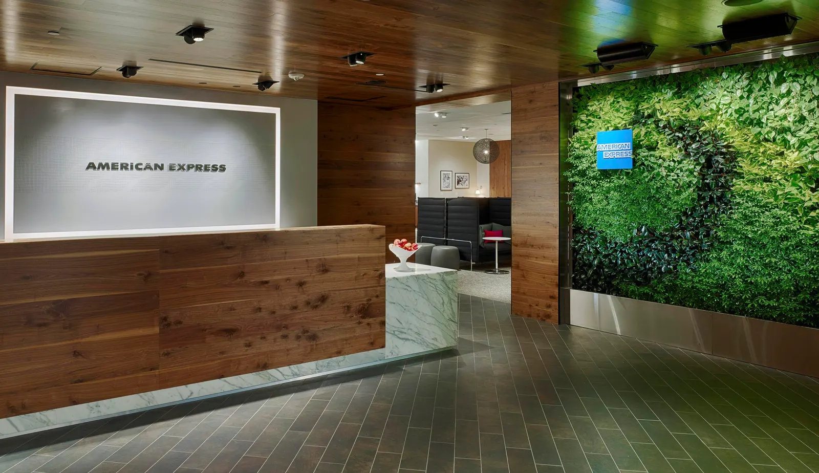 Wood-clad reception desk with a lit American Express logo panel and living green wall at the Centurion Lounge at Houston George Bush Intercontinental Terminal D (IAH).