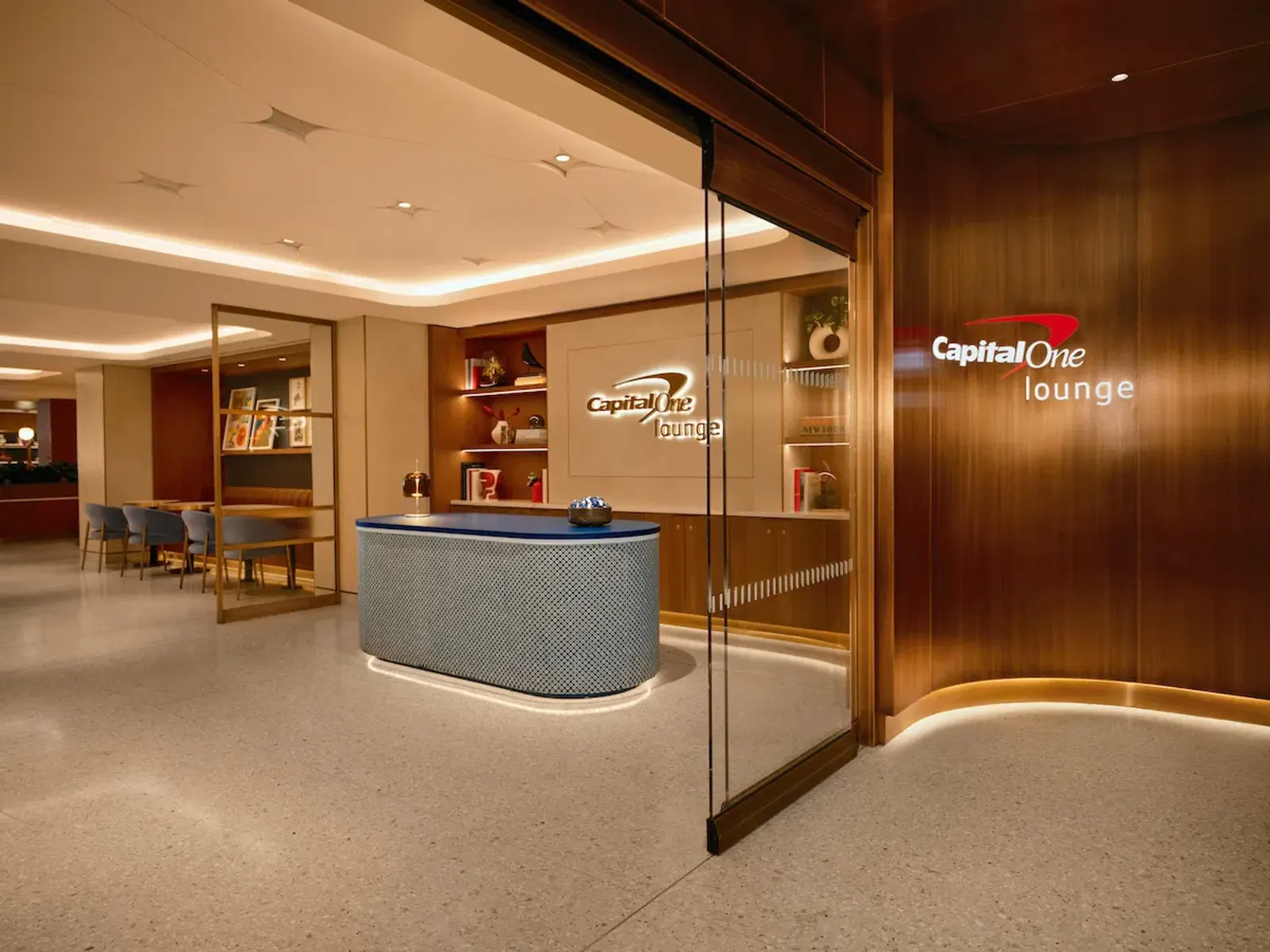 Curved wood-paneled entrance with Capital One lounge logo and blue reception desk at the Capital One Lounge at New York JFK Terminal 4.