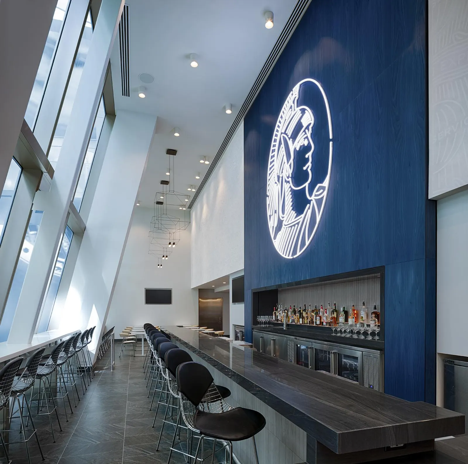Long wood bar with wire-frame stools under a glowing Centurion neon mark beside floor-to-ceiling windows in the Centurion Lounge at LAX.