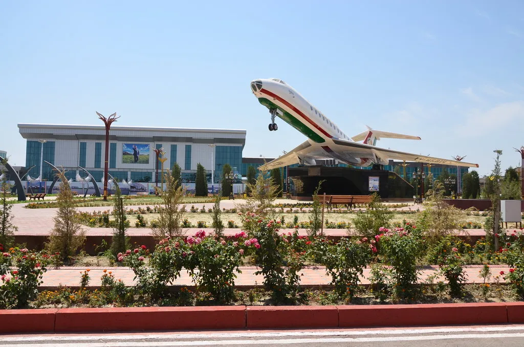Modern glass-fronted Khujand airport terminal behind a landscaped garden with rose beds and a retired airliner mounted on a plinth