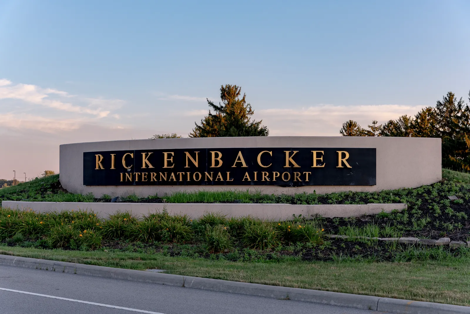 Entrance sign reading Rickenbacker International Airport on a planted berm beside the airport access road.