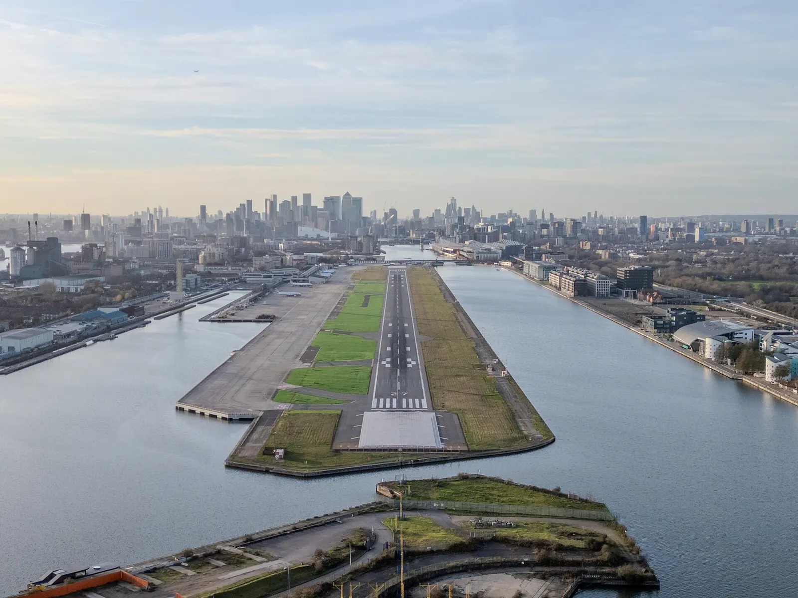 Aerial view west along London City Airport’s dock-island runway 09/27, with Canary Wharf and the City skyline beyond.