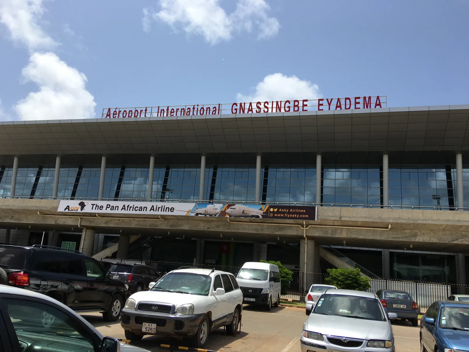 Front of the Gnassingbé Eyadéma International Airport terminal in Lomé, a long glass-and-steel building with the airport's name in red letters along the roof, with cars parked in the foreground under a blue sky